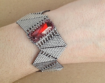 May include: A silver and red beaded bracelet with a geometric design. The bracelet features a large red gemstone in the center. The bracelet is called "Tangram bracelet".