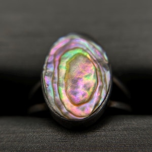 May include: An oval ring featuring a polished abalone shell inlay with iridescent swirls of pink, green, and purple. The shell is set within a dark metal band that has a smooth, rounded finish, highlighting the natural, multi-colored luster of the stone.