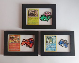 Puede incluir: Tres marcos de fotos negros con frentes de vidrio, cada uno contiene una carta de Pokémon y una versión de arte de píxeles del Pokémon. El marco superior presenta a Venusaur, el marco del medio presenta a Charizard y el marco inferior presenta a Blastoise.