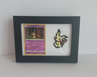 Puede incluir: Un marco negro con un paspartú blanco contiene una carta de Pokémon con Mimikyu, un Pokémon con un esquema de color amarillo y marrón. La carta está rodeada por un borde blanco y tiene el texto "Mimikyu -70" impreso en ella. El marco también contiene una imagen de pixel art de Mimikyu, un Pokémon con un esquema de color amarillo y marrón.