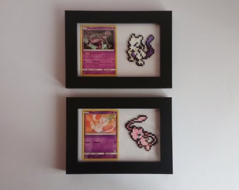 Puede incluir: Dos marcos de fotos negros con paspartús blancos. Cada marco contiene una carta de Pokémon y una versión de pixel art del Pokémon. El marco superior presenta una carta de Mewtwo y un Mewtwo de pixel art blanco. El marco inferior presenta una carta de Mew y un Mew de pixel art rosa.