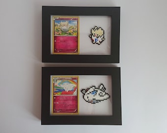 Puede incluir: Dos marcos de fotos negros con paspartús blancos. Cada marco contiene una carta de Pokémon y una versión de pixel art del Pokémon. El marco superior presenta una carta de Togepi y un Togepi de pixel art. El marco inferior presenta una carta de Togekiss y un Togekiss de pixel art.