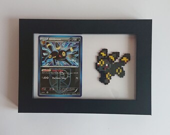 Puede incluir: Un marco negro con una carta de Pokemon que presenta a Umbreon, un Pokemon de tipo Oscuro con ojos amarillos. La carta está al lado de una versión de pixel art de Umbreon hecha con cuentas.