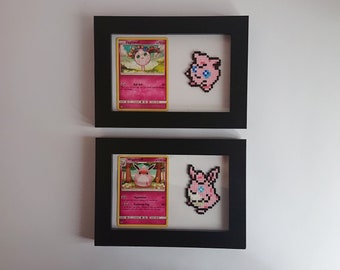 Puede incluir: Dos marcos de fotos negros con paspartús blancos, cada uno mostrando una carta de Pokémon y una versión de arte de píxeles del Pokémon. El marco superior presenta una carta de Wigglytuff y un Wigglytuff de arte de píxeles rosa. El marco inferior presenta una carta de Whimsicott y un Whimsicott de arte de píxeles rosa.