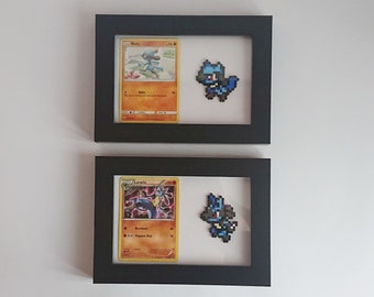 Puede incluir: Dos marcos de fotos negros con frentes de vidrio. Cada marco contiene una carta de Pokémon y una representación de pixel art del Pokémon. El marco superior presenta una carta de Riolu azul y blanca y un Riolu de pixel art azul. El marco inferior presenta una carta de Lucario azul y amarilla y un Lucario de pixel art azul.