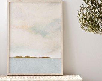 Puede incluir: Una pintura de acuarela enmarcada de un paisaje marítimo con un cielo azul claro y una línea de horizonte con una pequeña isla en la distancia. La pintura está colgada en una pared blanca sobre un baúl de almacenamiento de mimbre.