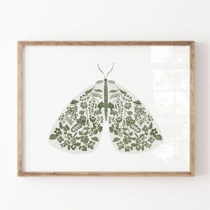 Green and white moth illustration with symmetrical wings filled with small flowers, leafy stems and botanical details, displayed in a simple natural wood frame on a white wall.