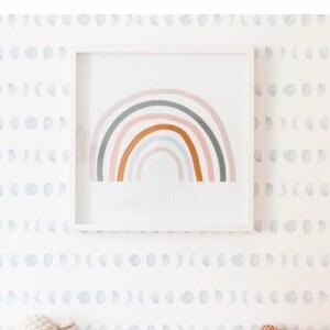 May include: A white cot with a white and grey moon phase wallpaper. A rainbow painting hangs above the cot. A stuffed lamb toy sits on the cot mattress. A brown and orange pillow sits on the cot.