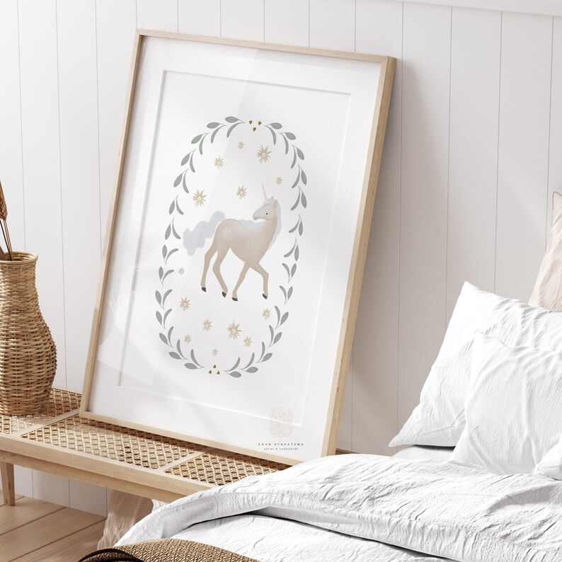 Framed unicorn wall art leaning on a wooden bench beside white bedding and a woven basket in a soft-lit bedroom.