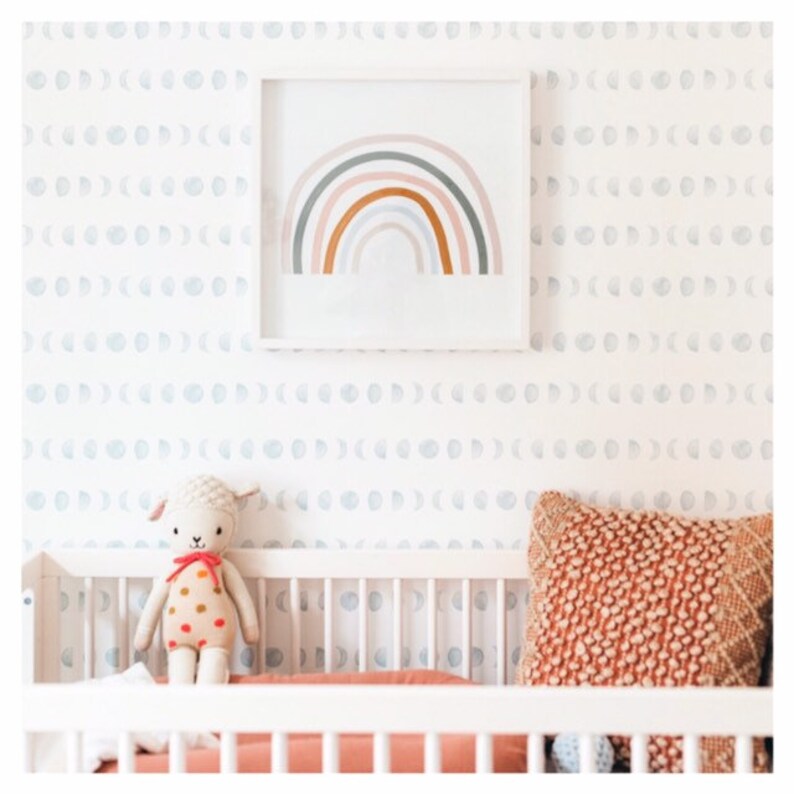 May include: A white cot with a white and grey moon phase wallpaper. A rainbow painting hangs above the cot. A stuffed lamb toy sits on the cot mattress. A brown and orange pillow sits on the cot.