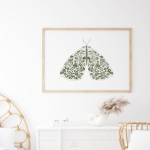 Framed floral moth artwork hanging above a white fireplace mantel, featuring pale wings decorated with green flowers and foliage in a bright neutral room.