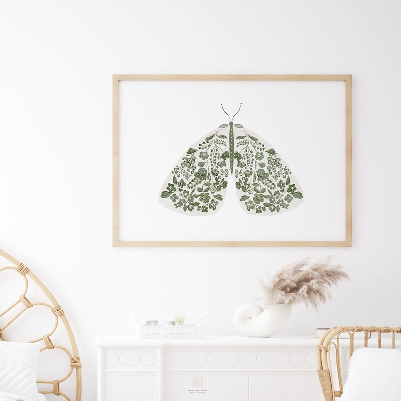 Framed floral moth artwork hanging above a white fireplace mantel, featuring pale wings decorated with green flowers and foliage in a bright neutral room.