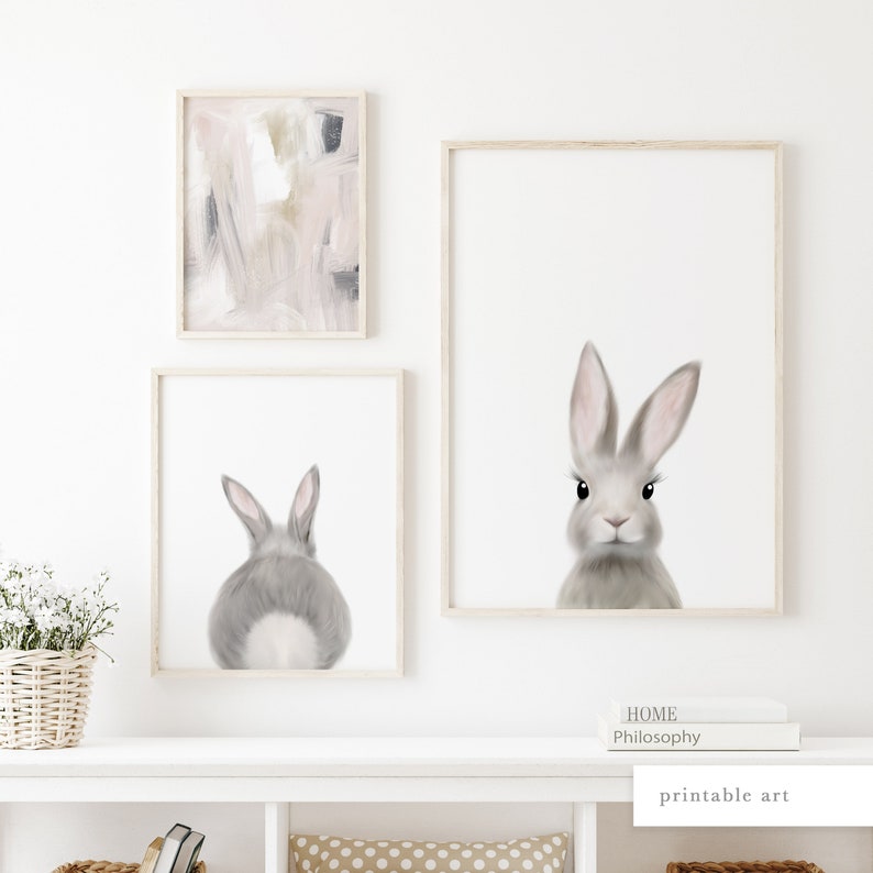 May include: Three framed prints on a white wall. The center print features a gray bunny rabbit with pink ears. The print on the left features a pink and gray abstract painting. The print on the right features a gray bunny rabbit looking at the camera.