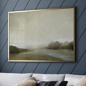 Framed moody landscape print with misty trees and a soft river, styled on a navy panelled wall above a neutral sofa with textured cushions.