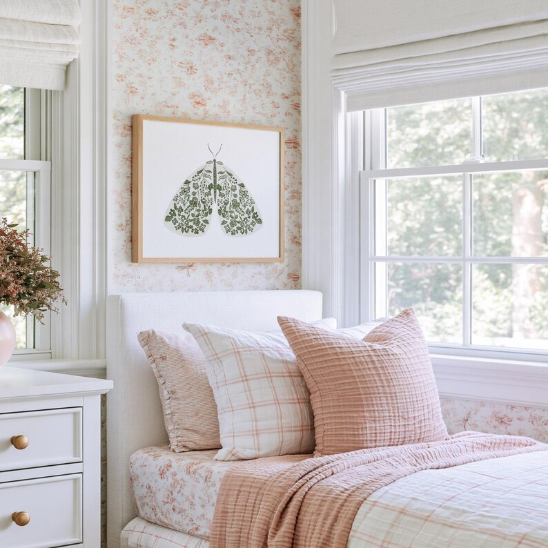 Green floral moth print in a light wood frame hanging above a bed in a bright bedroom with soft floral wallpaper, white bedding, blush pink pillows, a textured pink throw and large windows on either side.