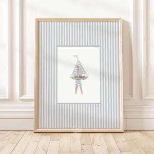 Nautical sailboat wall art in a blonde wood frame, leaning against a white paneled wall with light wood floors in a coastal nursery setting.