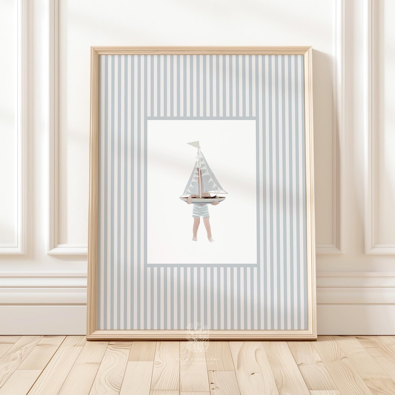 Nautical sailboat wall art in a blonde wood frame, leaning against a white paneled wall with light wood floors in a coastal nursery setting.