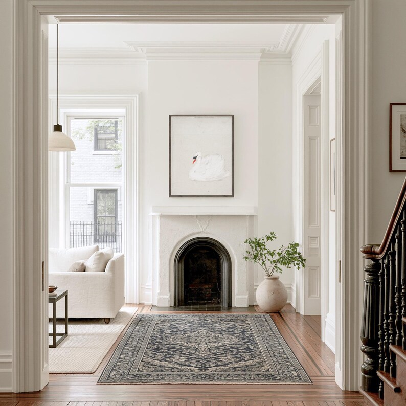 Vintage swan artwork displayed above a black fireplace at the end of a bright hallway, with white walls, wood floors, a patterned runner rug and a large ceramic vase nearby.