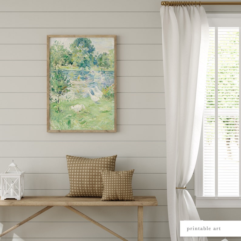 Puede incluir: Una pintura enmarcada que representa una escena de paisaje con un lago, &aacute;rboles y p&aacute;jaros blancos. La pintura est&aacute; colgada en una pared blanca con paneles de madera. Hay un banco de madera con dos cojines marrones delante de la pintura.