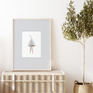Sailboat wall art in a light wood frame above a wicker hamper, paired with a potted olive tree in a softly styled kids room or entryway.