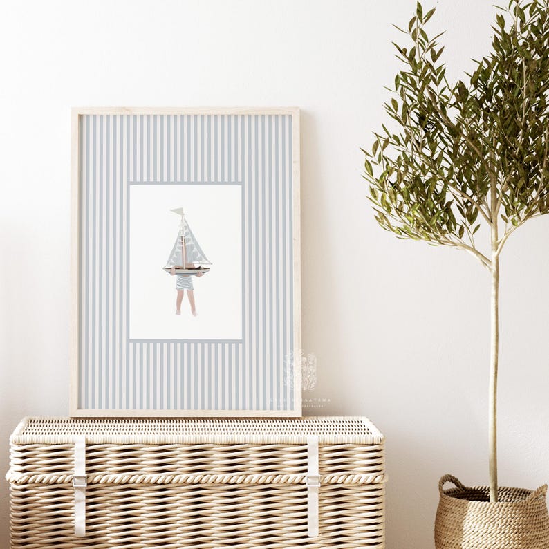 Sailboat wall art in a light wood frame above a wicker hamper, paired with a potted olive tree in a softly styled kids room or entryway.