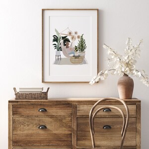 K&ouml;nnte beinhalten: Eine gerahmte Illustration einer Frau, die sich in einer H&auml;ngematte entspannt, umgeben von Pflanzen und Sonnenblumen. Die Illustration ist auf wei&szlig;em Hintergrund und in einem naturfarbenen Holzrahmen gerahmt. Der Rahmen h&auml;ngt an einer wei&szlig;en Wand &uuml;ber einer Kommode aus Holz mit Schubladen.