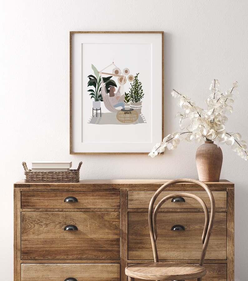 K&ouml;nnte beinhalten: Eine gerahmte Illustration einer Frau, die sich in einer H&auml;ngematte entspannt, umgeben von Pflanzen und Sonnenblumen. Die Illustration ist auf wei&szlig;em Hintergrund und in einem naturfarbenen Holzrahmen gerahmt. Der Rahmen h&auml;ngt an einer wei&szlig;en Wand &uuml;ber einer Kommode aus Holz mit Schubladen.