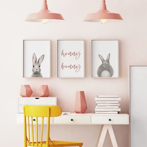 May include: Three framed prints with a gray bunny rabbit, the text "hunny bunny", and the back of a gray bunny rabbit. The prints are hanging on a light pink wall above a white desk with a yellow chair, a pink vase, and a stack of white books.