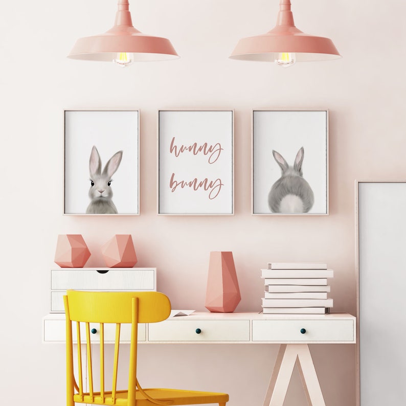 May include: Three framed prints with a gray bunny rabbit, the text "hunny bunny", and the back of a gray bunny rabbit. The prints are hanging on a light pink wall above a white desk with a yellow chair, a pink vase, and a stack of white books.