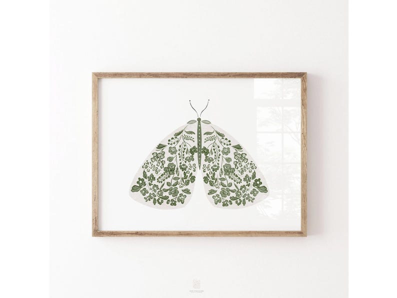 Green and white moth illustration with symmetrical wings filled with small flowers, leafy stems and botanical details, displayed in a simple natural wood frame on a white wall.