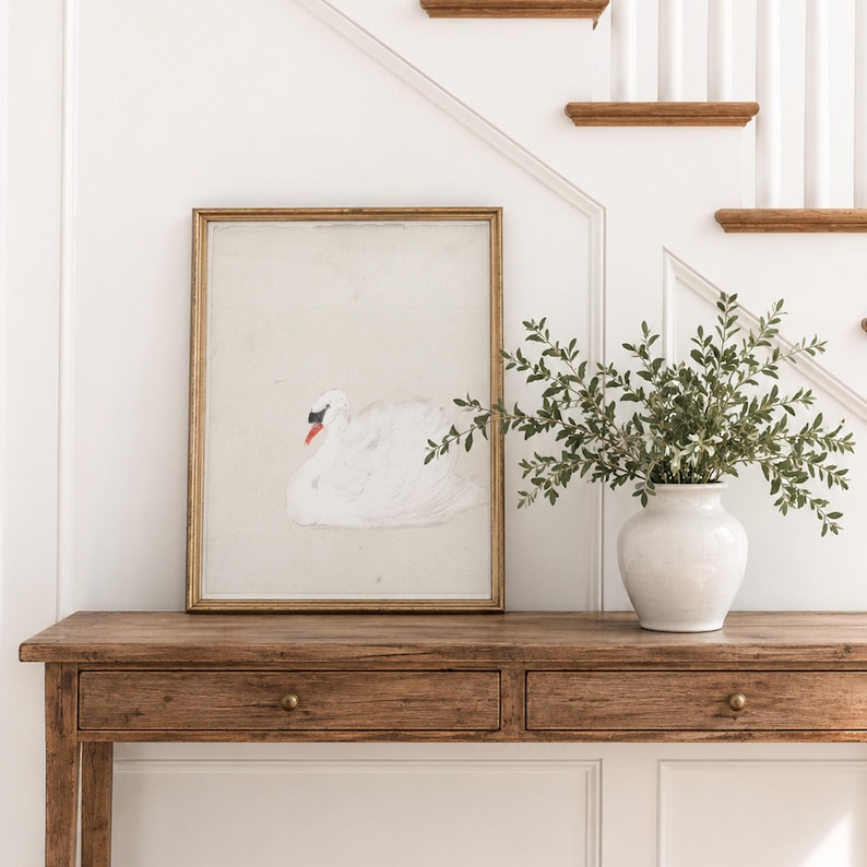 Framed vintage swan print resting on a rustic wood console table beside a white vase with leafy branches, featuring a white swan with a dark face and orange beak.