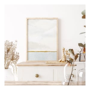 Elegant watercolour painting of a coastal horizon. The artwork showcases soft blue-grey skies merging with a serene blue sea, accented by a thin golden green shoreline, capturing a sense of calm and simplicity.
