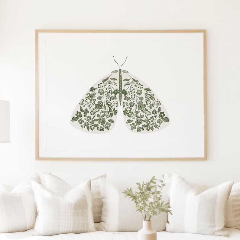 Large framed green moth illustration above a cream sofa, showing symmetrical white wings covered with delicate green flowers, leaves and stems in a light neutral living room.