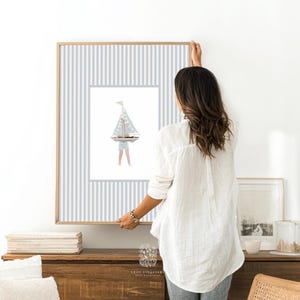 Sailboat art being hung above a wooden dresser in a clean, natural-toned space, perfect for styling in a boys room or lakehouse nursery.