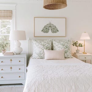 Green floral moth print in a thin wood frame centered above a white bed with floral pillows, flanked by white nightstands and lamps in a bright bedroom with a woven ceiling light and window.