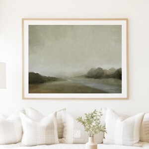 Framed moody nature print with misty trees and a soft river, styled above a white sofa with textured pillows and a minimalist ceramic vase.
