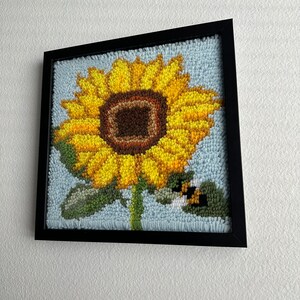 Könnte beinhalten: Ein gerahmtes Nadelbild mit einer gelben Sonnenblume mit brauner Mitte und grünem Stiel. Eine kleine Biene befindet sich in der Nähe der Sonnenblume.