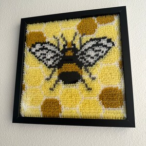 以下が含まれることがあります： 黄色と黒の蜂が黄色い蜂の巣の背景に描かれた、額縁に入った針仕事の作品です。