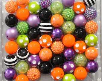 May include: A collection of round beads in a variety of finishes including solid orange, purple, black, and lime green, along with polka-dot patterns, black and white stripes, and textured glitter surfaces arranged in a grid on a clear tray.