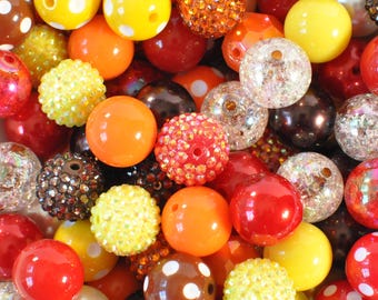 Könnte beinhalten: Eine Sammlung bunter Perlen in verschiedenen Größen und Texturen. Die Perlen sind hauptsächlich in Rot-, Orange-, Gelb-, Braun- und Klartönen gehalten. Einige Perlen haben Polka-Punkte, Glitzer oder eine strukturierte Oberfläche. Geeignet für Schmuckherstellung und Bastelarbeiten.