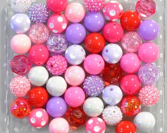 Könnte beinhalten: Eine Sammlung farbenfroher Perlen in verschiedenen Rosa-, Rot-, Lila- und Weißtönen. Einige Perlen sind einfarbig, während andere Glitzer, Tupfen oder facettierte Oberflächen aufweisen. Die Perlen sind in einem transparenten, quadratischen Behälter angeordnet.