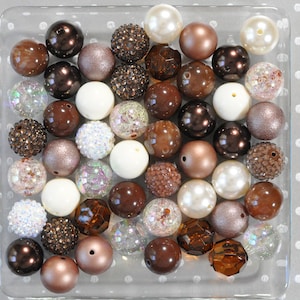 May include: A collection of brown, white, and iridescent beads in various textures and finishes. The beads are arranged in a clear glass dish.