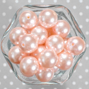 May include: A clear glass bowl filled with numerous light pink, round pearls. The pearls have a glossy, iridescent finish and a small hole drilled through each. The bowl sits on a gray surface with white polka dots.