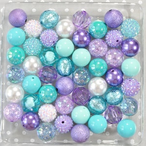 May include: A collection of various beads in shades of purple, teal, and white, displayed in a clear square container. The beads vary in size, texture, and finish, including pearlized, glitter, and faceted designs. The beads are arranged on a white dotted surface.
