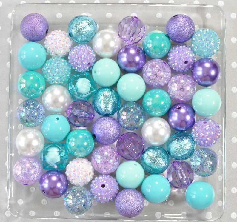 May include: A collection of various beads in shades of purple, teal, and white, displayed in a clear square container. The beads vary in size, texture, and finish, including pearlized, glitter, and faceted designs. The beads are arranged on a white dotted surface.