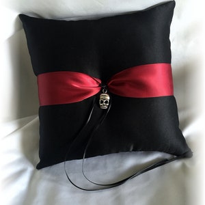 May include: Black satin pillow with a red satin ribbon and a silver skull charm. The pillow is for a wedding ceremony.