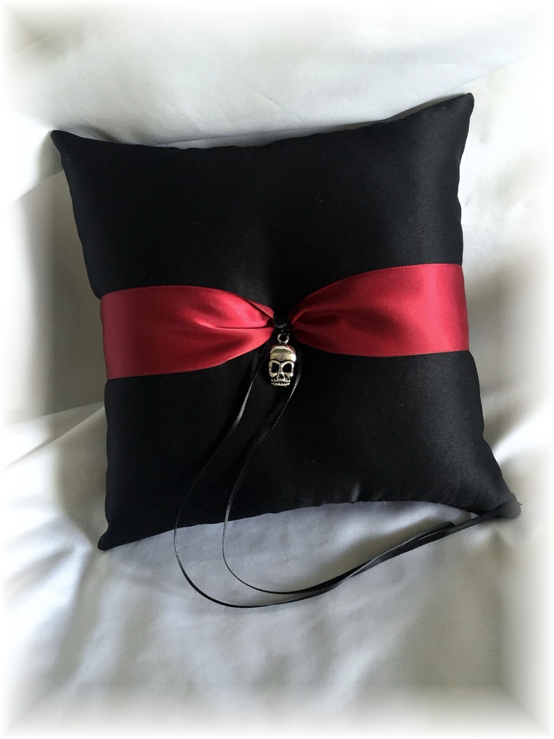 May include: Black satin pillow with a red satin ribbon and a silver skull charm. The pillow is for a wedding ceremony.