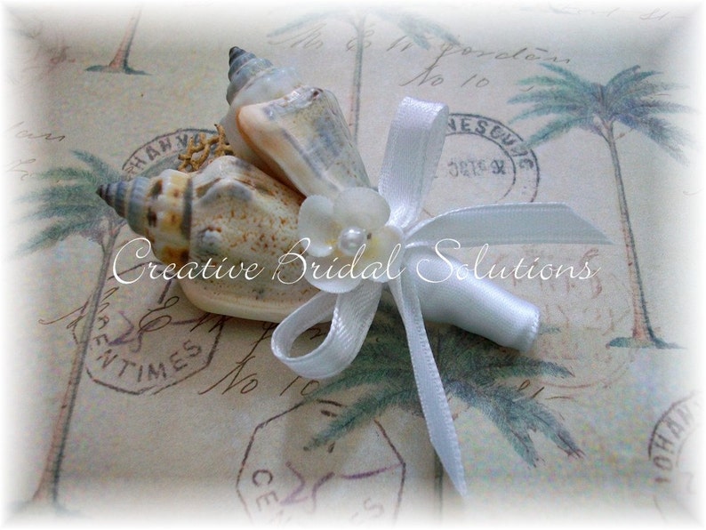 May include: A white satin ribbon tied around a bouquet of seashells. The bouquet is decorated with a white pearl and a white flower.