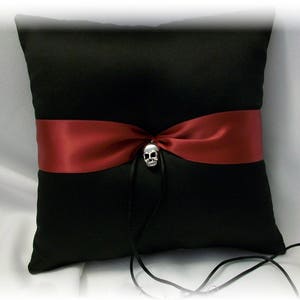 May include: Black satin pillow with a burgundy satin ribbon and a silver skull charm. The pillow is for a wedding ceremony.