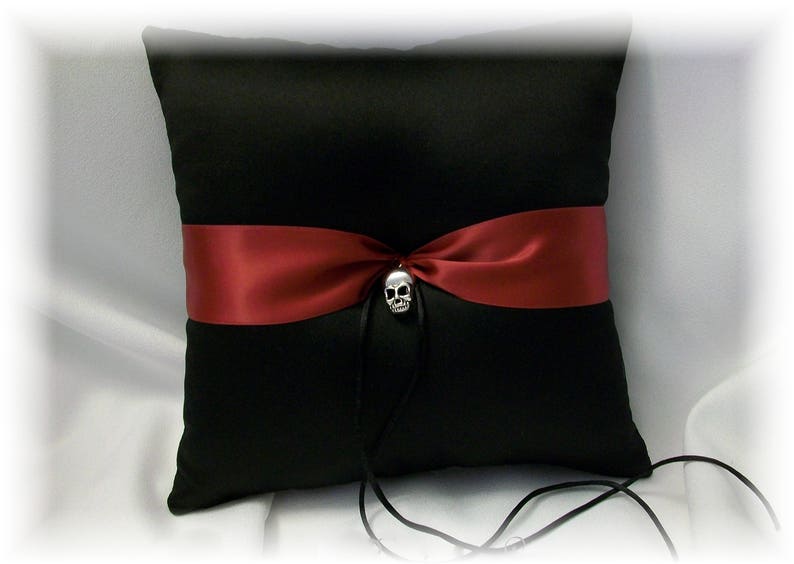 May include: Black satin pillow with a burgundy satin ribbon and a silver skull charm. The pillow is for a wedding ceremony.
